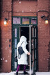 A woman in winter clothes opens the door to a building on the street during a snowfall.