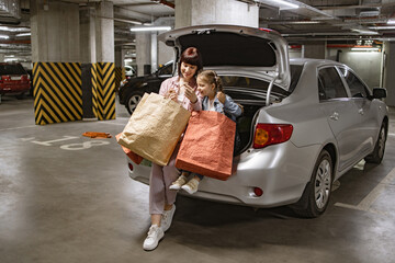 Dressed in casual attire Caucasian family mother and little daughter sitting on open trunk of car in underground parking lot, holding colorful bags with clothes from shopping mall.