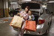 © sofiko14 - Adult female and child holding colorful bags with clothes purchased from shopping mall. Underground parking lot, Caucasian mother and small daughter sitting on open trunk of modern car.