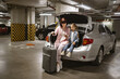 © sofiko14 - Caucasian mother and little daughter sitting in trunk of modern car in underground parking lot. Young woman with child holding traveling suitcase using phone for planning vacation trip.