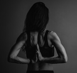 Wall Mural - Female sport muscular woman doing stretching workout the shoulders, blades and arms in sport black bra, standing on dark shadow background with empty copy space foe text. Back view. Black and white