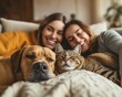 Two women are laying on a bed, one holding a dog while the other cuddles a cat. The room is cozy and relaxed.