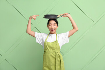 Poster - Portrait of young Asian woman pointing at frying pans on her head against green background