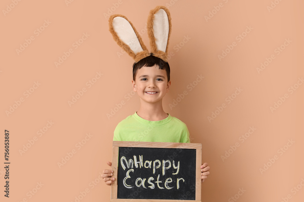 Cute little boy in bunny ears holding chalkboard with text HAPPY EASTER on beige background