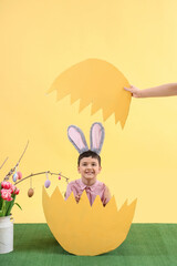 Poster - Cute little boy with bunny ears in paper egg shell on yellow background. Easter celebration