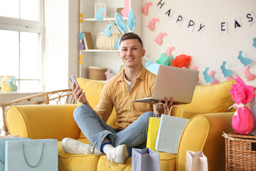 Wall Mural - Happy young man in bunny ears with mobile phone and laptop sitting at home. Easter Sale