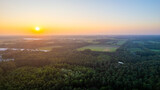 Fototapeta Natura - This image captures the serene beauty of a sunset horizon viewed from above a verdant forest. The setting sun dips towards the horizon, its warm hues painting the sky in gradients of orange, yellow