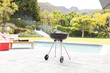 A charcoal grill is ready for a barbecue by a poolside with lush greenery in the background