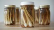 Jars of pickled garleek strands, preserved in a clear brine with mustard seeds and peppercorns, neatly lined up against a soft backlit background, evoking a home-canning charm.