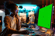 © Gorodenkoff - Portrait of Creative Black Woman Sitting at Her Desk Using Desktop Computer with Mock-up Green Screen. Female Specialist Working at Night in Game Development Startup, Finishing Details on a Project
