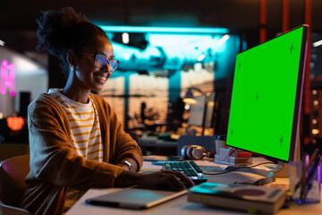 Fototapete - Portrait of Creative Black Woman Sitting at Her Desk Using Desktop Computer with Mock-up Green Screen. Female Specialist Working at Night in Game Development Startup, Finishing Details on a Project