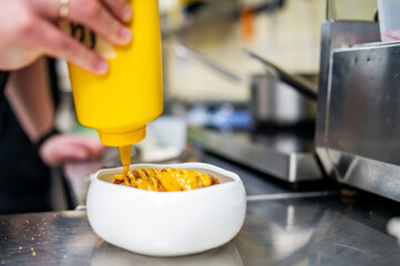 Wall Mural - Chef in a professional kitchen drizzling mustard from a yellow bottle onto a dish, with blurred kitchen equipment in the background.