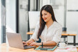 © PRIME STOCK LAB - Smiling asian businesswoman in a casual white blouse sits confidently at her desk with a computer in a bright, modern office.
