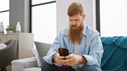 Wall Mural - Focused young redhead man, chilling on his sofa indoors, engrossed in texting on his smartphone at his cozy apartment.