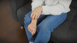 © Krakenimages.com - A young woman in casual attire scratching her arm while sitting on a gray couch indoors.