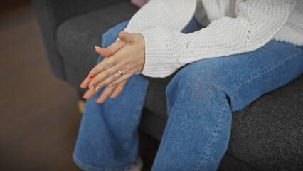 Wall Mural - A woman in a white sweater and blue jeans sits indoors, hands clasped on her lap, depicting casual home relaxation.