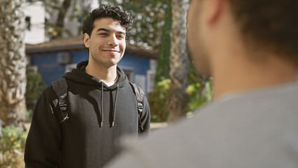 Sticker - Two hispanic men smiling and chatting on a sunny park pathway, exuding friendship and leisure in an urban outdoor setting.