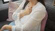 Middle-aged woman clutching chest in discomfort while seated on a sofa indoors, conveying potential health issues.