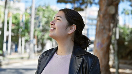 Smiling young hispanic woman in casual attire enjoying the outdoors in a city park with trees and daylight.