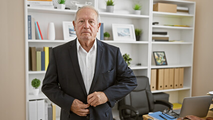 Sticker - Serious-faced, senior businessman standing confidently in an office interior, working successfully as a seasoned worker. wearing an elegant jacket, focused and relaxed on his job.