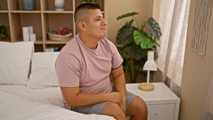 Canvas Print - Serious young latin man sitting in cozy bed, concentrating deeply in his comfortable apartment's bedroom, presenting a handsome expression in the quiet morning