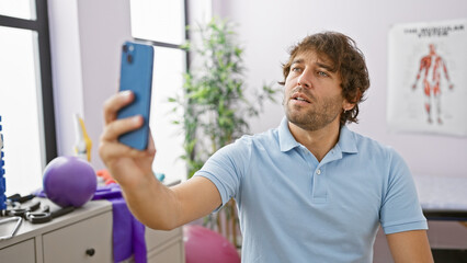 Canvas Print - A young caucasian man with a beard takes a selfie in a clinical room, representing healthcare professionals and social media use at work.