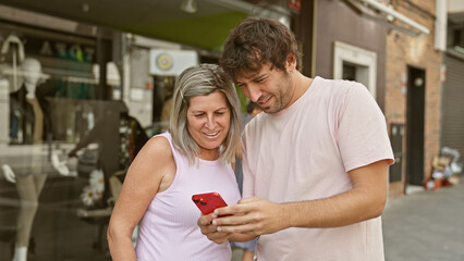 Poster - Casual city joy, confident caucasian mother and son standing together on an urban road, happily texting on a smartphone, their lovely smiles radiating absolute happiness in the embracing sunlight.