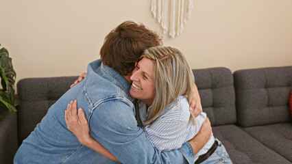 Wall Mural - Heartwarming mother and son sitting together on the living room sofa, joyfully hugging and smiling in their cozy home, radiating happiness and confidence in their casual lifestyle.