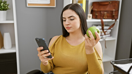 Wall Mural - Hispanic woman multitasking with smartphone and apple in modern office setting, exuding professionalism and health-consciousness.