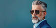 A sophisticated portrait of a mature man with silver hair and a neatly trimmed beard, wearing sunglasses and a sharp blue suit, against a solid blue background with copy space.