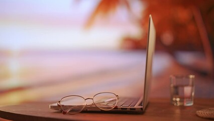 Close up shot of computer at the table. Closed laptop on the table with glasses and glass of water against tropical beach seaside background.