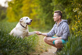 Fototapeta Londyn - Happy man with dog in nature on sunny summer day. Cute yellow labrador retriever giving paw to his pet owner..