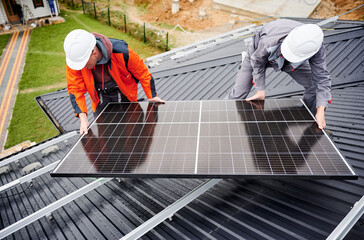 Wall Mural - Electricians installing solar panel system on roof of house. Men workers in helmets carrying photovoltaic solar module outdoors. Concept of alternative and renewable energy.