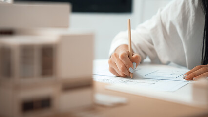 Closeup of beautiful young architect engineer hand drawing, drafting blueprint with blueprint and house model placed on working table at modern office. Focus on hand. Blurring background. Immaculate.