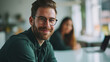© MP Studio - Young man with glasses in the foreground, smiling towards the camera, with coworkers in the background, in a meeting at office