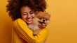 Portrait of smiling young woman with afro hairstyle in yellow sweater posing with cute Pomeranian spitz puppy. Happy girl hugging her beloved pet. Love between human and dog. Yellow studio background.