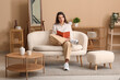 © Pixel-Shot - Beautiful young happy woman with book sitting on comfortable sofa in living room at home