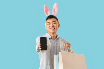 Wall Mural - Young Asian man in bunny ears with mobile phone and shopping bags on blue background. Easter Sale