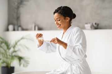 Wall Mural - Happy black woman in bathrobe preparing to brush her teeth, applying toothpaste