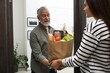 © New Africa - Courier giving paper bag with food products to senior man indoors