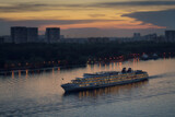 Fototapeta Natura - Ship is sailing on river near cityscape at golden sunset with reflections on the water in the city