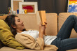 © Chanakon - Asian beautiful woman lay down on cozey couch sofa reading book in living room with warm light at night. People female stay home part time relax at home.