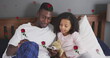 Image of roses over happy african american father and daughter reading book in bed