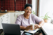 © shurkin_son - Happy cheerful retired african american plus size female in glasses holding cup of coffee sitting at kitchen near window in front of laptop, browsing internet and websites with culinary recipes