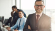 © Flamingo Images - Confident handsome young businessman stands in a boardroom, surrounded by diverse colleagues. He has his arms crossed and looks directly into the camera with a big smile