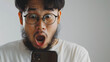 Close up snapshot of a 23-year-old asian man with beard in white shirt wearing glasses, with surprised look at cellphone display