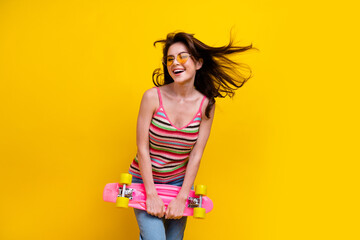 Poster - Portrait of lovely girl with fluttering hair wear striped tank in glasses hold skateboard isolated on bright yellow color background