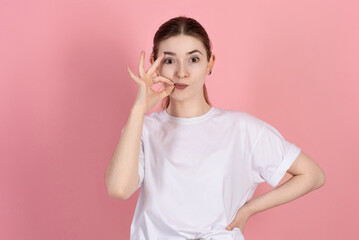 Wall Mural - Attractive caucasian young brunette woman in casual white t-shirt keeps secrets and shows gesture zip isolated on pink studio background.