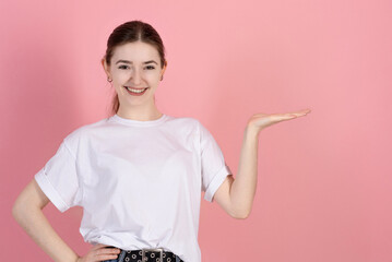 Wall Mural - Attractive Caucasian young brunette woman in casual white t-shirt shows palm empty copy space for product advertisement isolated on pink studio background.