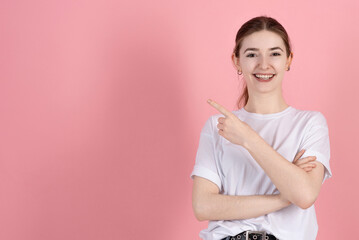 Wall Mural - Attractive Caucasian young brunette woman in casual white t-shirt pointing finger at empty copy space for advertisement isolated on pink studio background.